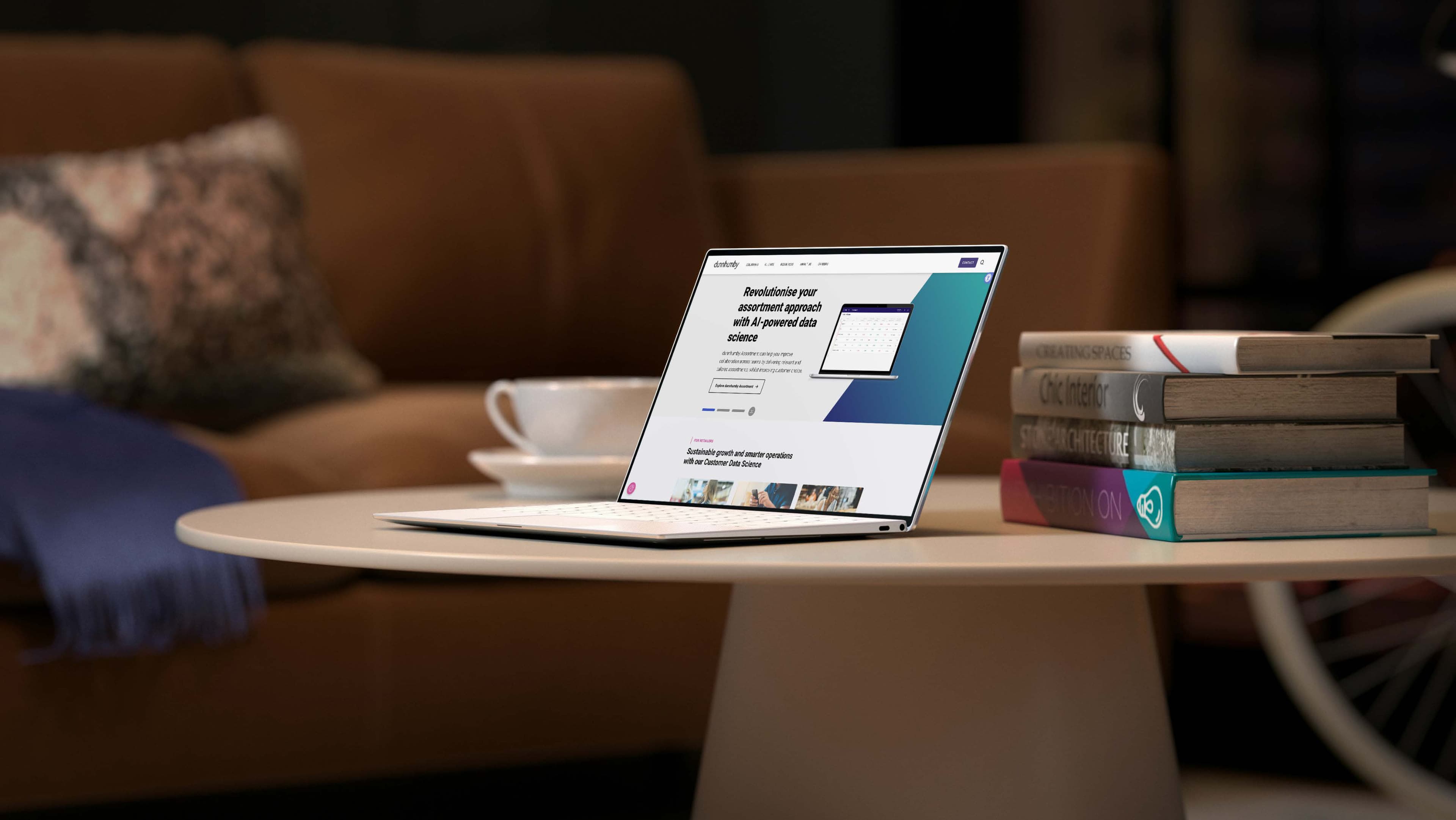 Laptop on a round white table displaying a website, next to a stack of books and a coffee cup in a cozy living room setting.